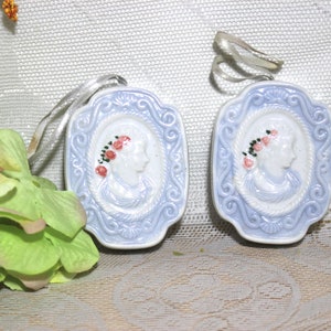 May include: Two blue and white porcelain ornaments with a raised design of a woman's profile with a floral crown. The ornaments have a rope-like border and a decorative scroll design.