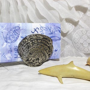 May include: A decorative seashell-shaped object with a textured surface, resting on a blue and white patterned background. A yellow shark figurine and seashells are also present. The background includes the word "MADE".