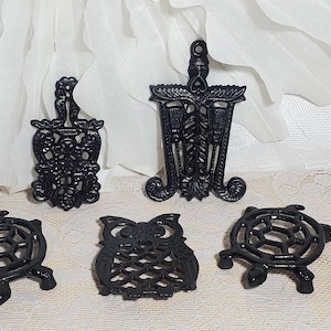 May include: A collection of five black cast iron trivets in various shapes. The trivets include a turtle, an owl, and decorative designs. These trivets are designed to protect surfaces from hot cookware.