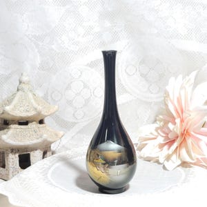 May include: A slender black vase with a narrow neck and a rounded base, featuring a decorative gold and silver landscape illustration on the front that depicts a mountain, trees, and architectural structures in a detailed, painted style.