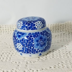 May include: A blue and white ceramic ginger jar with a lid. The jar is decorated with floral patterns in varying shades of blue. The lid also features a floral design. The jar sits on a white surface.
