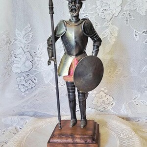 May include: A silver-toned figurine of a knight in armor, holding a spear and shield. The figure stands on a wooden base, with details including a hat and facial features. The background is a white lace fabric.