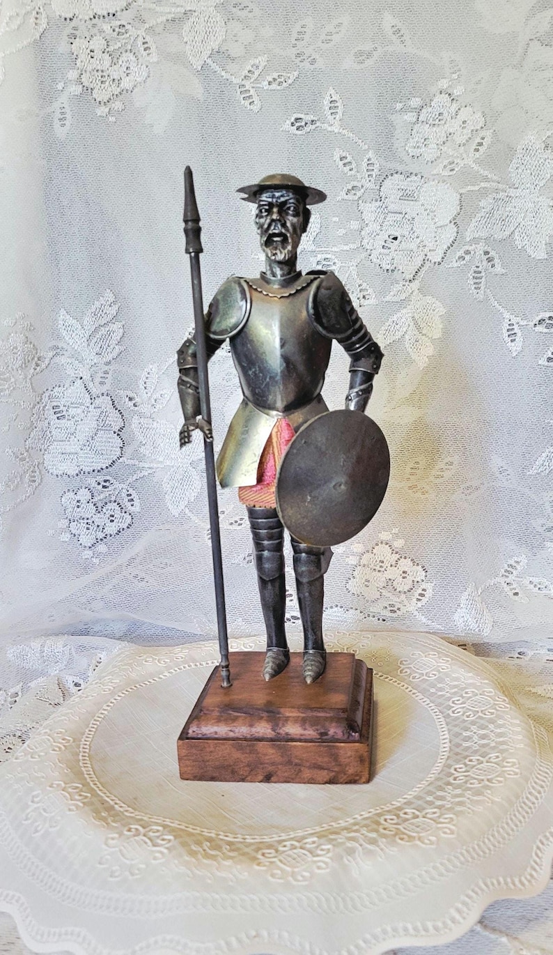 May include: A silver-toned figurine of a knight in armor, holding a spear and shield. The figure stands on a wooden base, with details including a hat and facial features. The background is a white lace fabric.