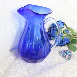May include: A cobalt blue glass pitcher with a clear handle. The pitcher has a ruffled rim and a ribbed body. A small glass heart and blue flowers are also in the image.