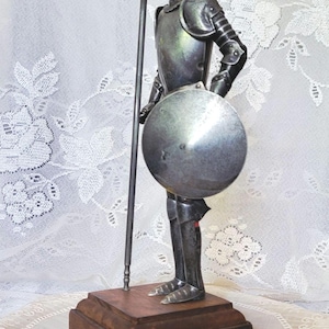 May include: A metal figurine of a knight in armor, holding a spear and shield. The figure stands on a wooden base. The knight wears a helmet and is posed in a stance of readiness. The figurine is silver in color.