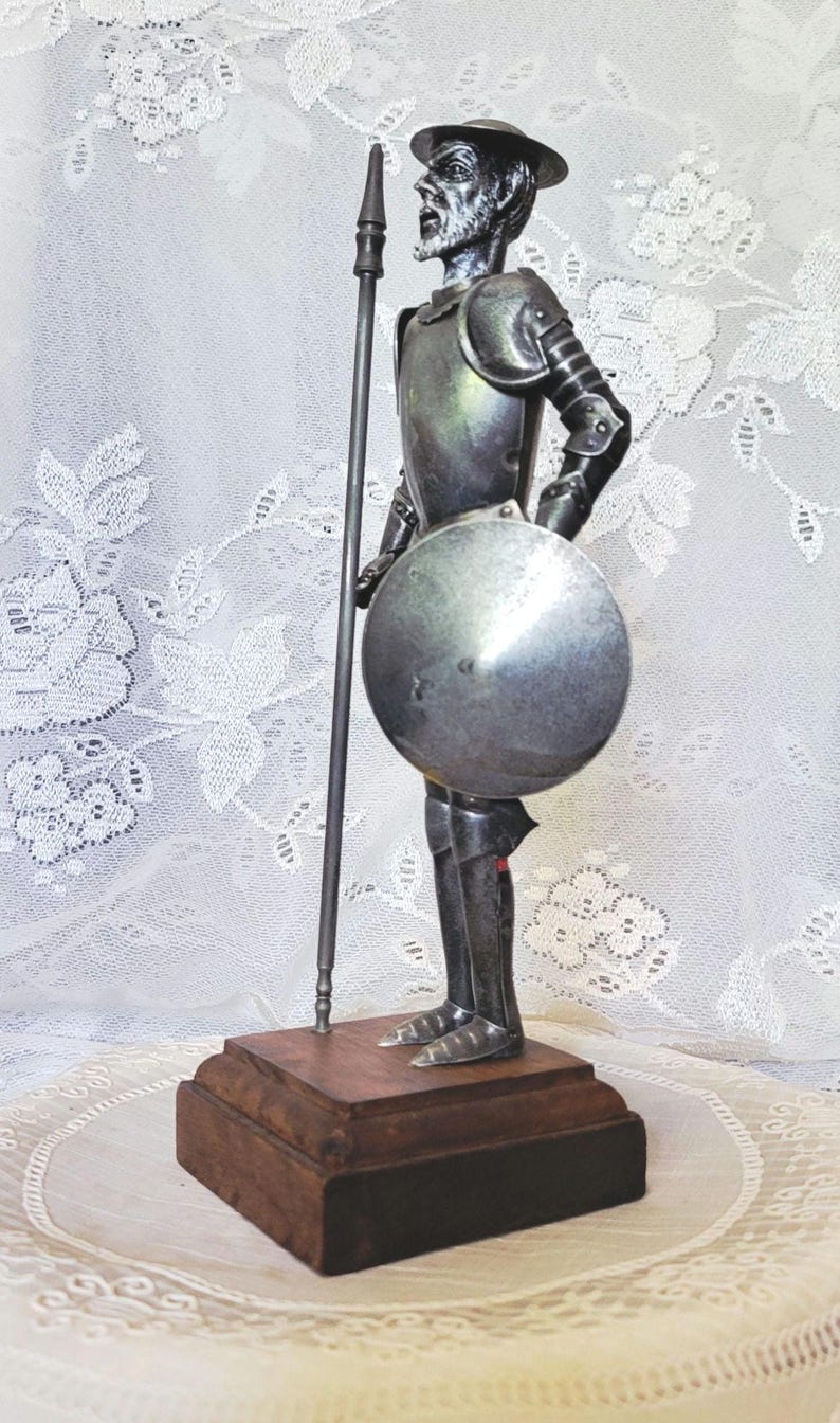 May include: A metal figurine of a knight in armor, holding a spear and shield. The figure stands on a wooden base. The knight wears a helmet and is posed in a stance of readiness. The figurine is silver in color.