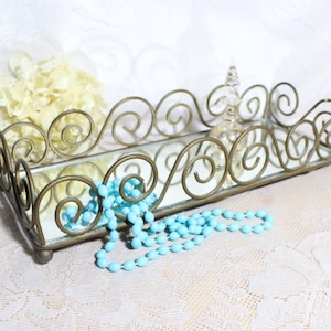May include: A gold-toned metal tray with a mirrored surface and a decorative scroll design. The tray is filled with a turquoise blue beaded necklace.