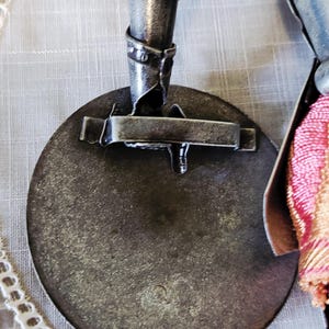 May include: Close-up of a metal decorative object with a circular base and a mechanical arm. The object has a distressed, aged appearance with visible welds and rivets. A decorative tassel with pink and gold threads is attached.