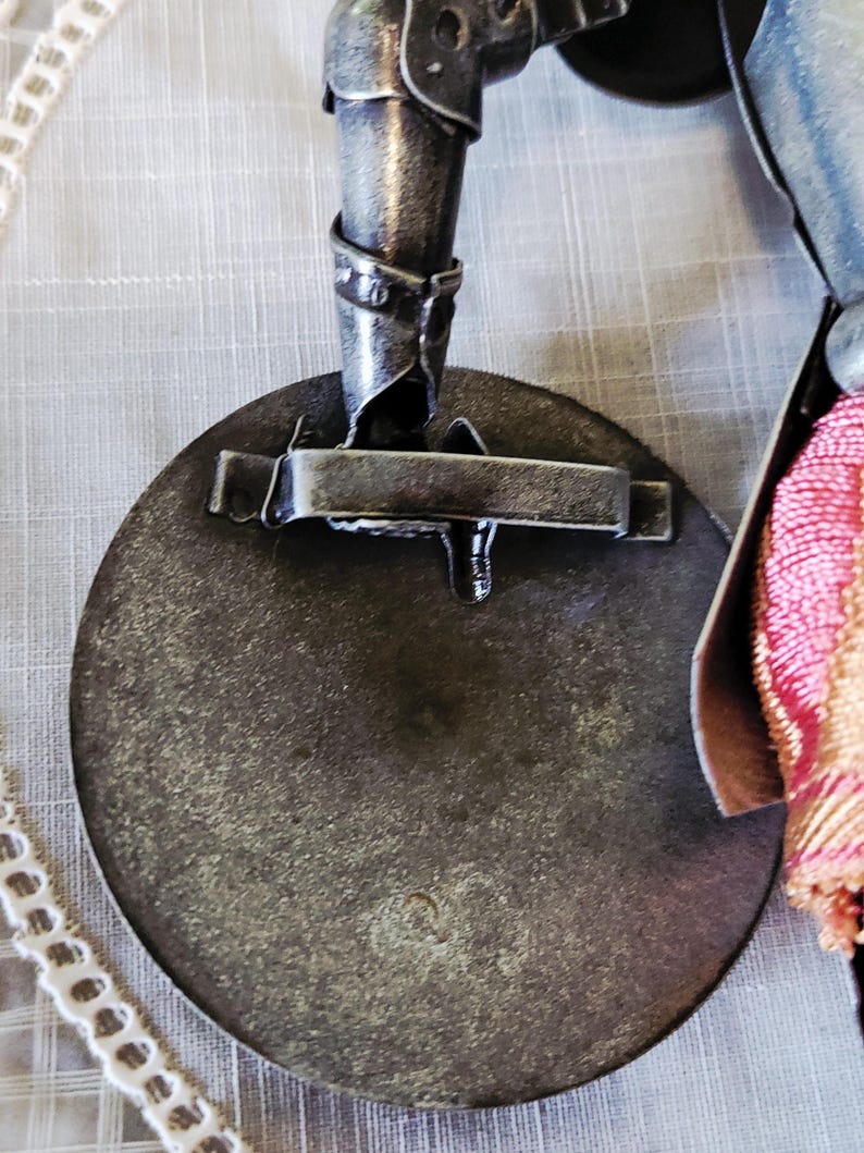 May include: Close-up of a metal decorative object with a circular base and a mechanical arm. The object has a distressed, aged appearance with visible welds and rivets. A decorative tassel with pink and gold threads is attached.