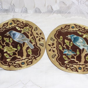 May include: Two round, decorative pieces with a brown background and gold trim. Each features a blue bird perched on a branch with green leaves and flowers. The design is intricate and detailed.