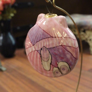 May include: A pink ceramic ornament shaped like a seashell with a gold top. The ornament has a painted design of pink, purple, and white swirls and lines.