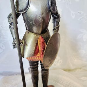 May include: A silver-toned figurine of a knight in full armor, holding a spear and a shield. The knight's armor has intricate details, and a red and gold fabric is visible beneath the armor. The figurine is mounted on a wooden base.