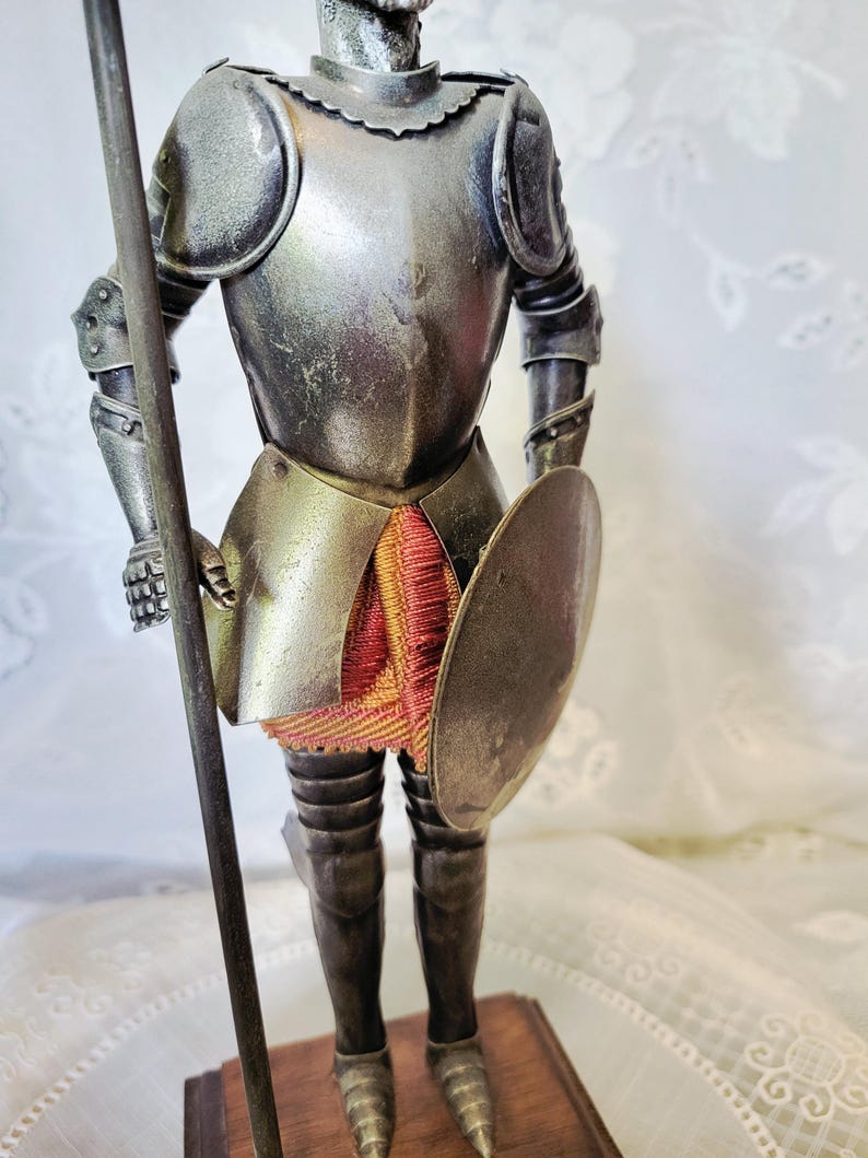 May include: A silver-toned figurine of a knight in full armor, holding a spear and a shield. The knight's armor has intricate details, and a red and gold fabric is visible beneath the armor. The figurine is mounted on a wooden base.