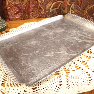 May include: A rectangular, silver-colored metal tray with embossed floral patterns. The tray has a rolled edge on one side and a scalloped edge on the other. It rests on a white, crocheted doily, suggesting a vintage or antique aesthetic.