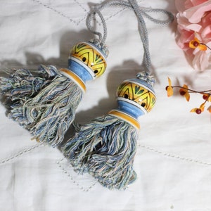 May include: Two decorative tassel tiebacks. Each tassel features a ceramic top with yellow, blue, and green patterns, and a blue and yellow base. The tassels are blue, green, and white. The tiebacks are on a white surface.