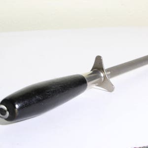 Puede incluir: Un afilador de cuchillos de acero con un mango de madera negro. El afilador tiene una varilla larga y cil&iacute;ndrica de acero y un protector cerca del mango. El mango es oscuro y liso, con una tapa met&aacute;lica y un peque&ntilde;o lazo.