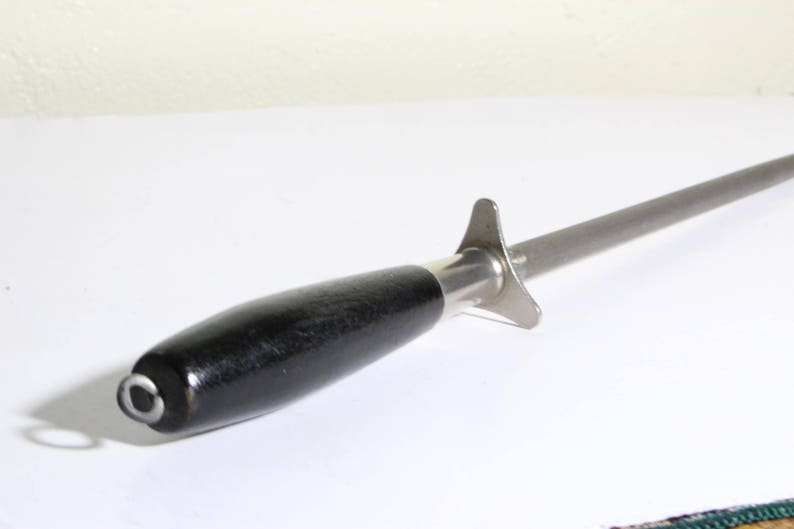 Puede incluir: Un afilador de cuchillos de acero con un mango de madera negro. El afilador tiene una varilla larga y cil&iacute;ndrica de acero y un protector cerca del mango. El mango es oscuro y liso, con una tapa met&aacute;lica y un peque&ntilde;o lazo.
