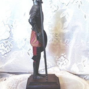 May include: A metal figurine of a knight in armor, holding a spear and shield. The knight is standing on a wooden base. The armor is dark gray, and the knight wears a red and gold sash. The figurine is a decorative item.