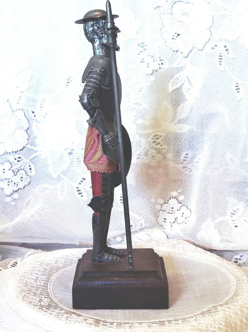 May include: A metal figurine of a knight in armor, holding a spear and shield. The knight is standing on a wooden base. The armor is dark gray, and the knight wears a red and gold sash. The figurine is a decorative item.