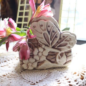 May include: A ceramic butterfly-shaped vase with a floral design, holding pink and yellow flowers. The vase is off-white with brown accents. The flowers have pink petals and green stems.