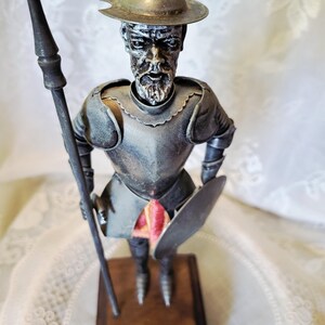 May include: A metal figurine of a knight, holding a spear and shield. The knight wears a helmet and armor, and stands on a wooden base. The figurine has a weathered appearance, with a touch of red fabric visible.