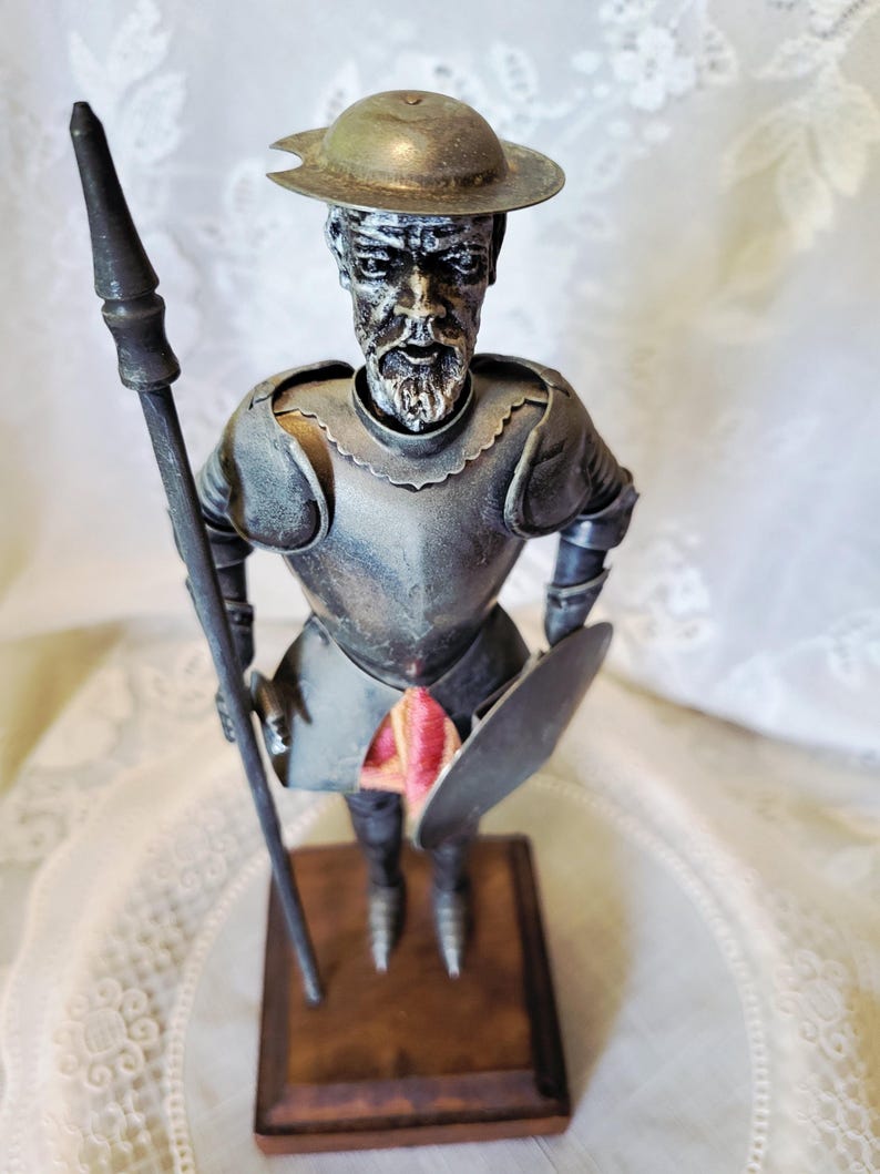 May include: A metal figurine of a knight, holding a spear and shield. The knight wears a helmet and armor, and stands on a wooden base. The figurine has a weathered appearance, with a touch of red fabric visible.