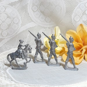 May include: Four silver toy soldiers marching in a line, with one soldier on horseback. The soldiers are holding rifles and wearing hats. The soldiers are standing on a white surface with a yellow flower behind them.