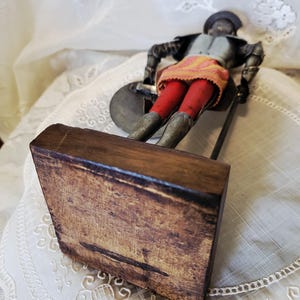 May include: A vintage figurine of a soldier in armor, holding a shield and a spear. The figure wears a gray helmet, a gray and black jacket, red pants, and a yellow and orange skirt. The figurine is mounted on a wooden base.