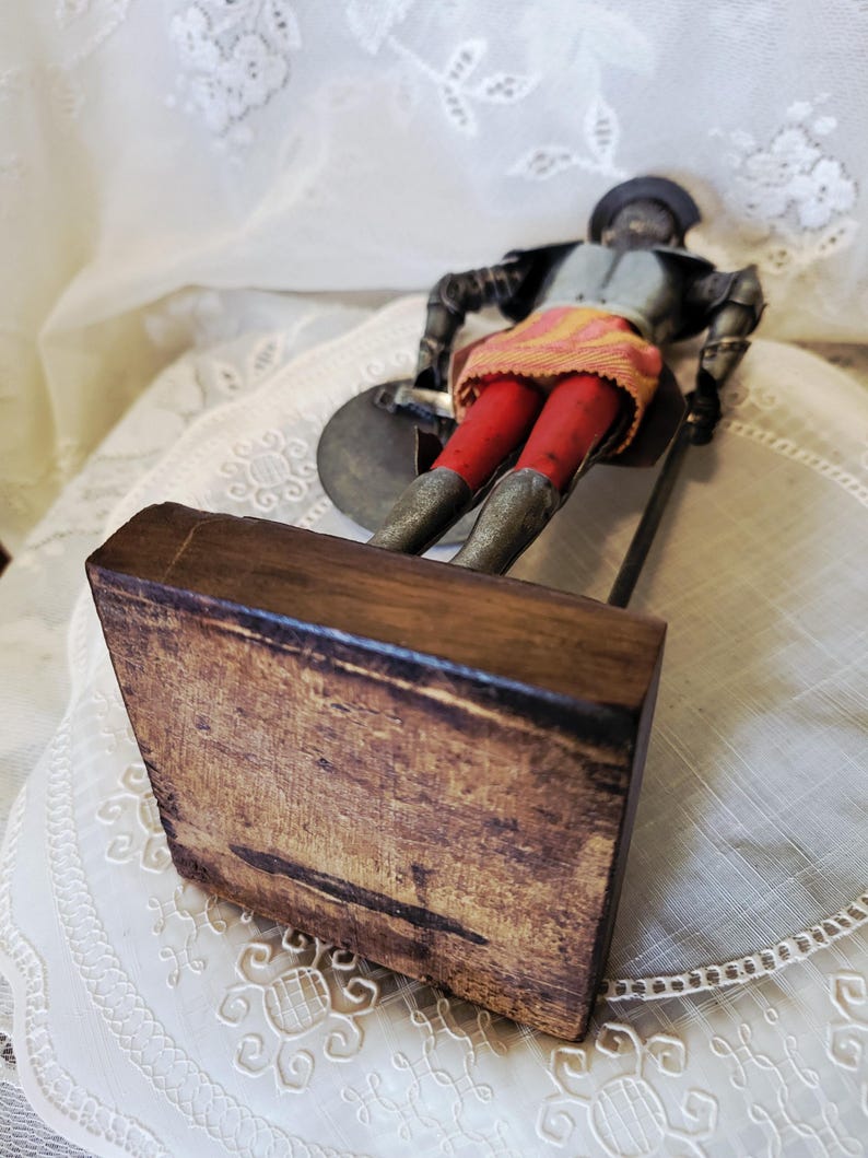 May include: A vintage figurine of a soldier in armor, holding a shield and a spear. The figure wears a gray helmet, a gray and black jacket, red pants, and a yellow and orange skirt. The figurine is mounted on a wooden base.