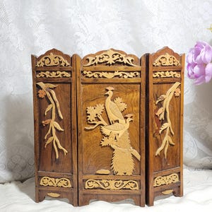 May include: A three-panel wooden room divider with intricate carvings. The center panel features a carved design of peacocks on a branch. The wood is a dark brown with gold accents.