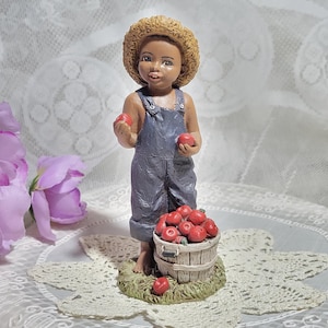 May include: A figurine of a young boy wearing overalls and a straw hat, holding red apples and standing next to a basket full of red apples.