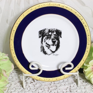 May include: Decorative plate with a black and white dog portrait. The plate has a white center, a navy blue border, and a gold rim. It is displayed on a white metal stand. The plate is a decorative item.
