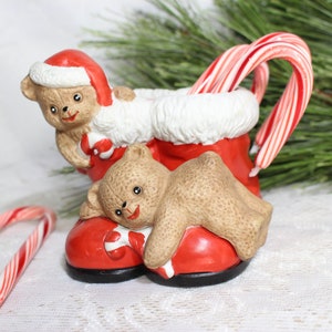 May include: A ceramic Christmas decoration featuring a red boot with a white fur trim, holding two candy canes. Two teddy bears are part of the design, one peeking over the top and the other resting on the boot. A candy cane lies on the surface.
