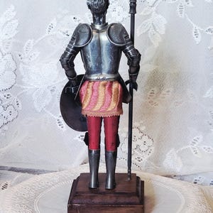 May include: A metal figurine of a knight in armor, holding a spear and shield. The knight wears a silver helmet, armor, and boots, with red pants and a pink and gold skirt. The figurine is mounted on a wooden base.