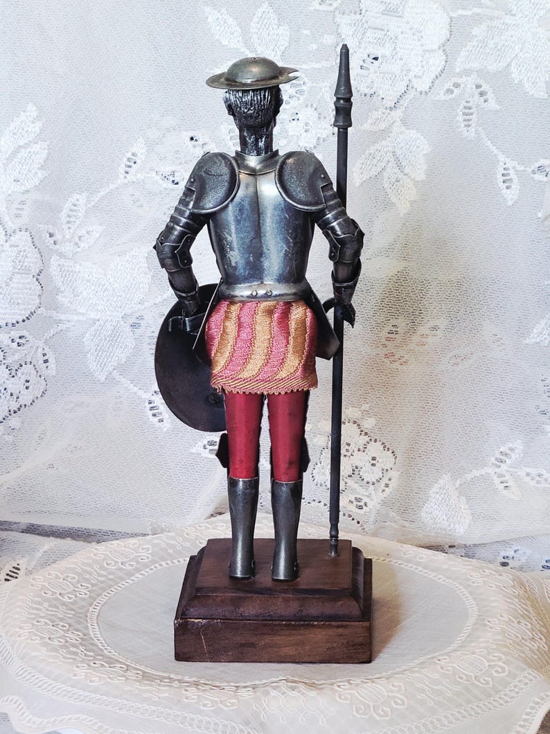 May include: A metal figurine of a knight in armor, holding a spear and shield. The knight wears a silver helmet, armor, and boots, with red pants and a pink and gold skirt. The figurine is mounted on a wooden base.