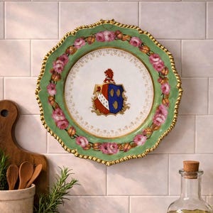 Könnte beinhalten: Ein dekorativer Teller mit einem grünen, gewellten Rand und goldenem Rand. Der Teller hat ein zentrales Wappenmotiv und ist von rosa Rosen umgeben. Der Teller hängt an einer weißen gefliesten Wand.