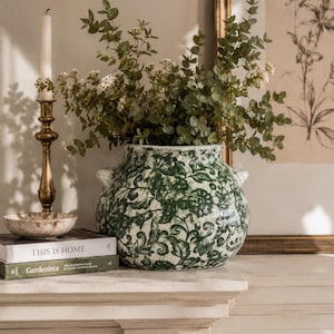 Puede incluir: Un jarrón de cerámica redondo con base blanca y un patrón floral verde oscuro, que cuenta con dos pequeñas asas laterales. El jarrón está lleno de ramas de eucalipto verdes y pequeñas flores blancas, colocado junto a libros y un candelabro de latón.