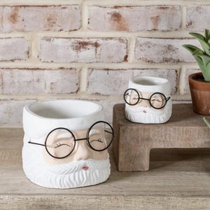 May include: Two white ceramic planters shaped like a face with a white beard, black round glasses, and a pink nose and mouth. The larger planter is on a wooden surface, and the smaller one is on a wooden block. The background is a white brick wall.