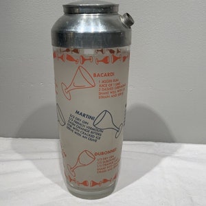 May include: A vintage silver cocktail shaker with a glass bottom. The glass is printed with cocktail recipes for Bacardi, Martini, and Dubonnet. The recipes are printed in blue and orange text.