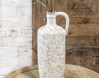 May include: A white ceramic vase with a handle and a floral pattern in shades of beige and brown. The vase is cylindrical with a slightly rounded base and a narrow neck. It contains several thin, dried stems.