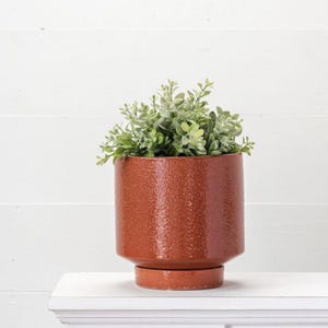 May include: A terracotta-coloured ceramic planter with a textured surface, holding a cluster of green leafy plants. The planter has a cylindrical shape with a small, round base. The plant pot is approximately 18 cm in diameter and 15 cm tall.