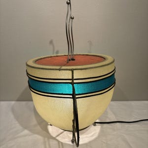 May include: A mid-century modern lamp with a cream-colored shade, a turquoise stripe, and a red interior. The lamp has a metal frame and a decorative metal element extending upwards. The lamp is on a white surface.