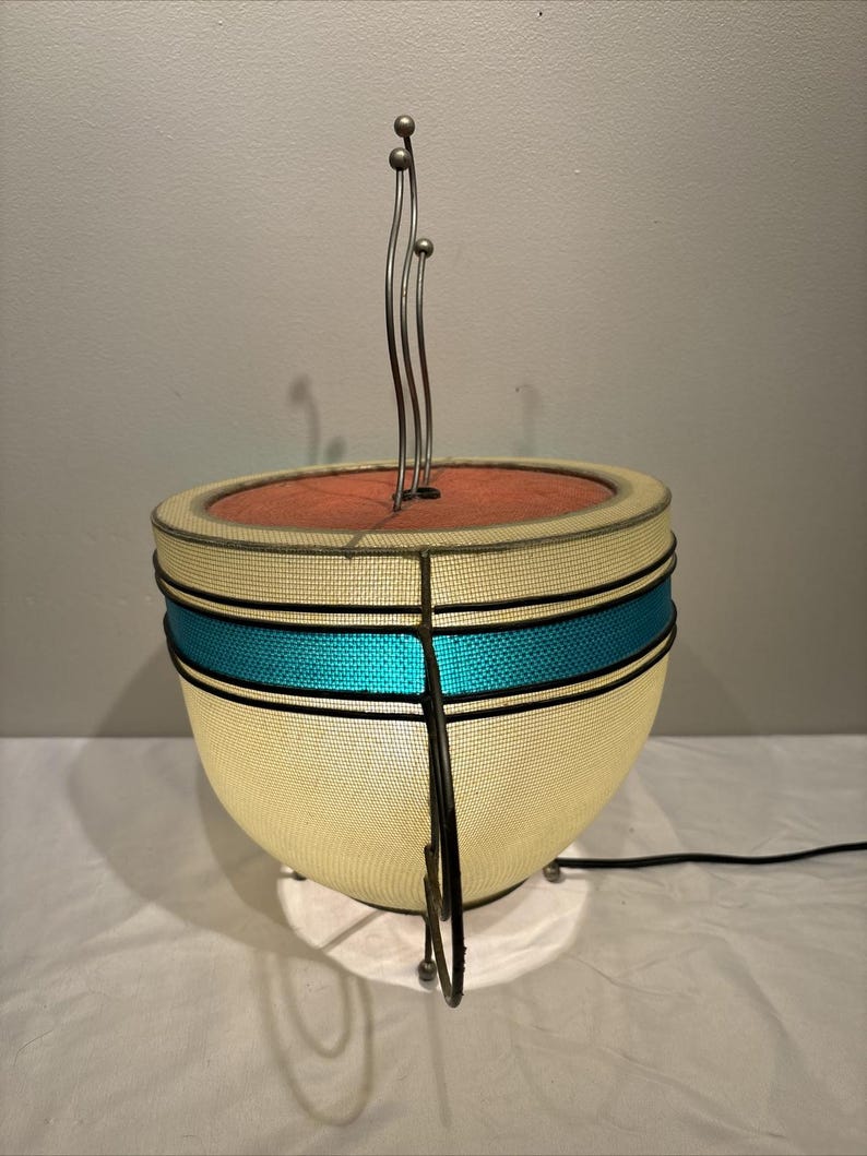May include: A mid-century modern lamp with a cream-colored shade, a turquoise stripe, and a red interior. The lamp has a metal frame and a decorative metal element extending upwards. The lamp is on a white surface.