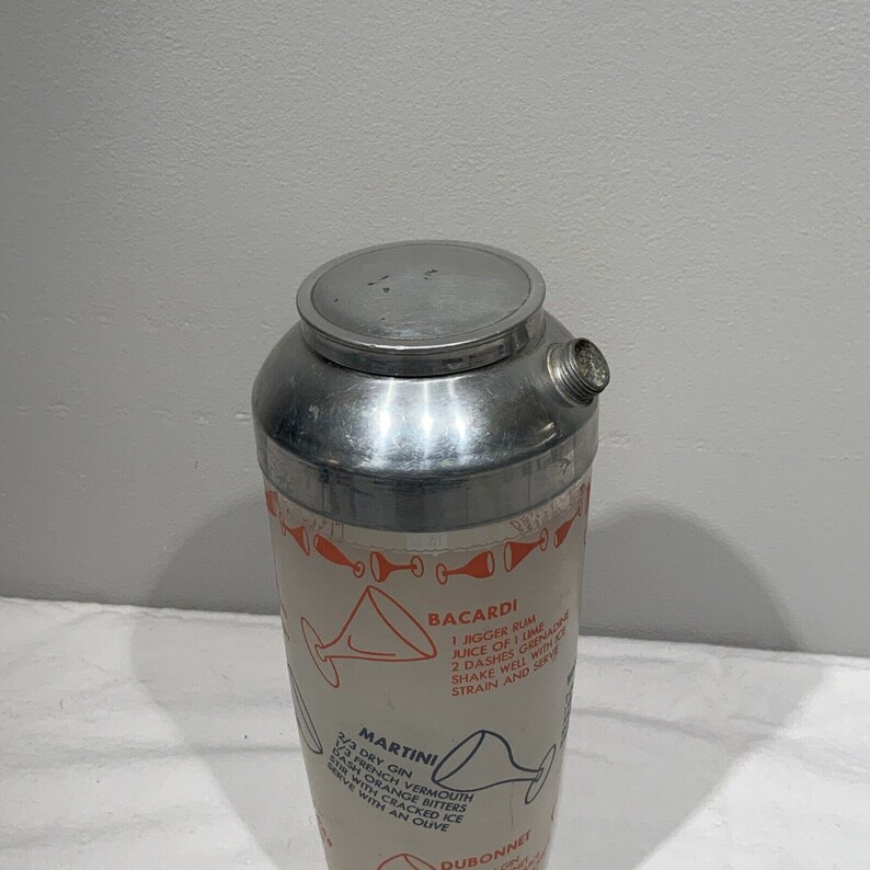 May include: A silver cocktail shaker with a white label and orange text. The label includes instructions for making a Bacardi, Martini, and Dubonnet cocktail.
