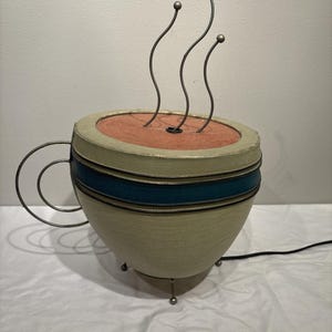 May include: A whimsical lamp shaped like a coffee cup. The cup is beige with a teal stripe and a wire handle. Three curved metal rods with ball tips rise from the cup's interior, simulating steam. The lamp has a black power cord.