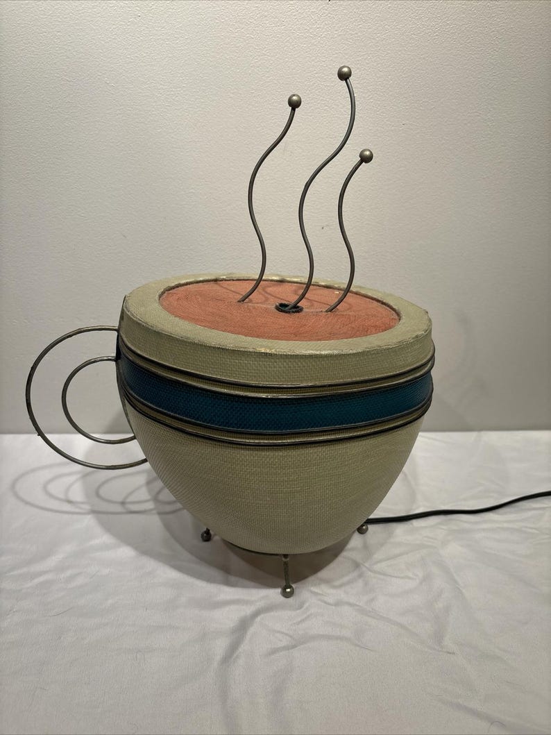 May include: A whimsical lamp shaped like a coffee cup. The cup is beige with a teal stripe and a wire handle. Three curved metal rods with ball tips rise from the cup's interior, simulating steam. The lamp has a black power cord.
