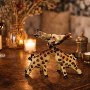 以下が含まれることがあります： 木製のテーブルの上に置かれた、水玉模様のセラミックキリンの置物。置物はキスをしているポーズです。背景にはワイングラス、ワインボトル、キャンドルがあります。