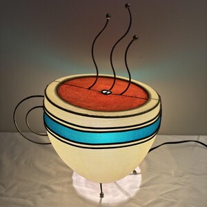 May include: A whimsical coffee cup lamp with a cream-colored fabric shade, accented by a vibrant blue stripe and a red interior. The lamp features a black metal handle and three curved metal accents that resemble steam. It stands on three small legs.