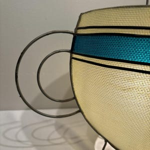 May include: A vintage-style hanging lamp with a cream-colored mesh shade and a teal blue band. The lamp features a metal frame with a circular handle design and three small feet. The lamp is lit from within.