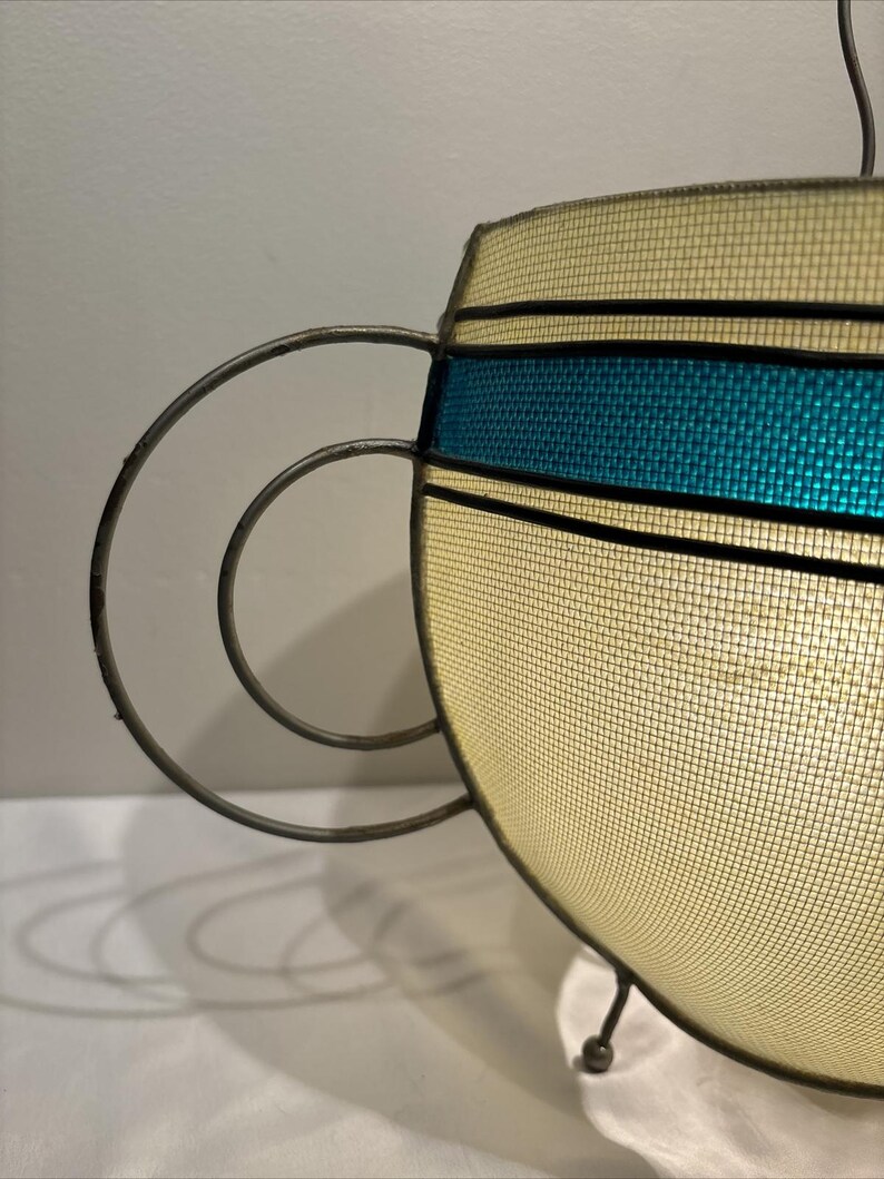 May include: A vintage-style hanging lamp with a cream-colored mesh shade and a teal blue band. The lamp features a metal frame with a circular handle design and three small feet. The lamp is lit from within.